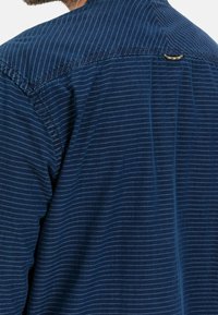 Tummansininen paita, jossa on vaakaraitoja, kaulus ja pieni kultainen yksityiskohta takana. Pehmeä materiaali ja rento muotoilu.