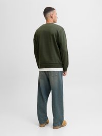 Mand vendt væk iført en olivengrøn sweatshirt, løse blå jeans, beige arbejdstøvler og en hvid skjorte under.