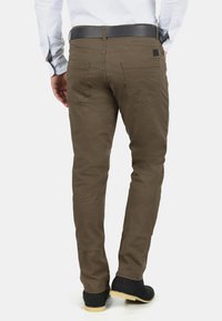 Pantalones marrones ajustados de algodón con una textura suave, que cuentan con bolsillos traseros y un cinturón negro, combinados con zapatos negros.