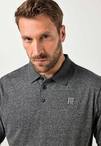 Uomo con capelli corti castani e barba, che indossa una polo grigio scuro dal tessuto strutturato con bottoni neri e un logo di simbolo matematico bianco sul petto.