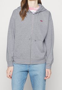 Sudadera gris con cremallera, con un bolsillo frontal, capucha con cordones y un pequeño logo rojo en el lado izquierdo del pecho. Fabricada en una suave mezcla de algodón texturizado.
