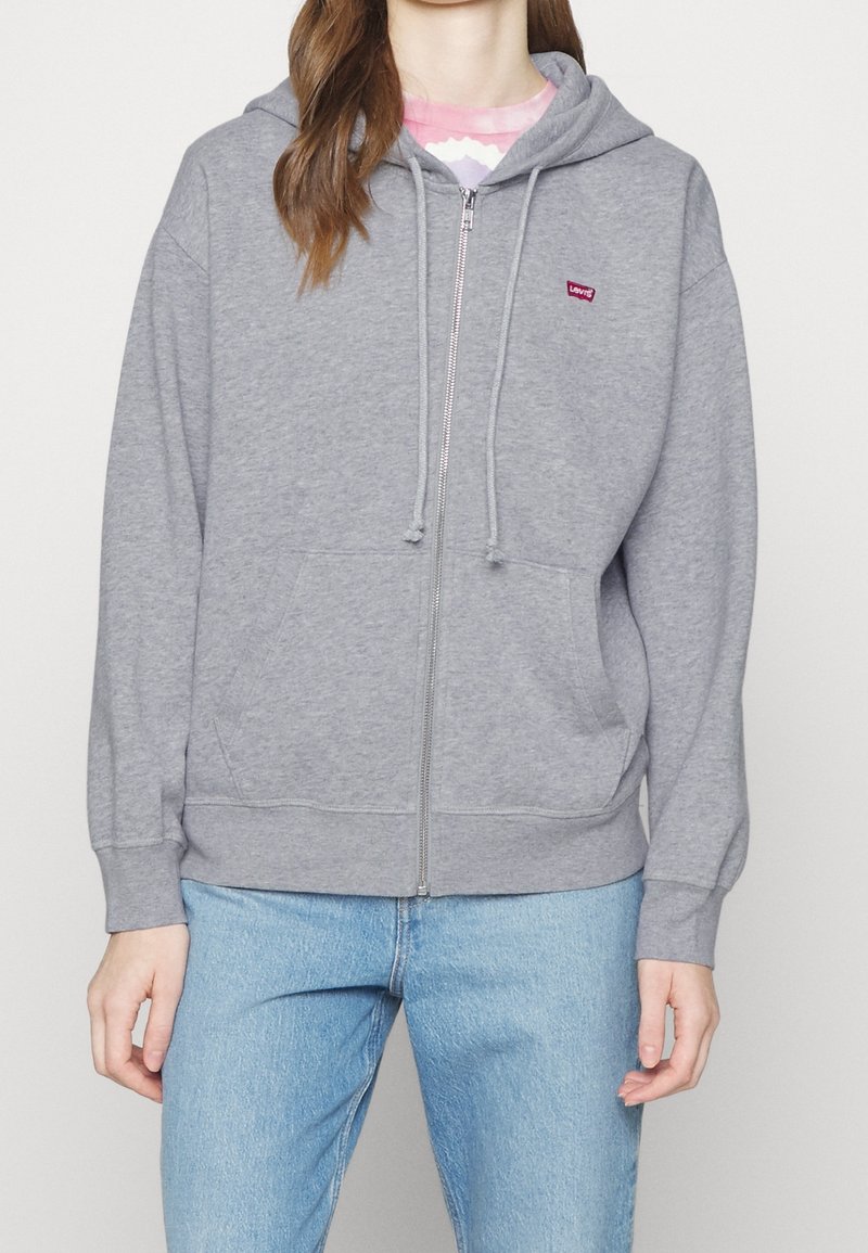 Sudadera gris con cremallera, con un bolsillo frontal, capucha con cordones y un pequeño logo rojo en el lado izquierdo del pecho. Fabricada en una suave mezcla de algodón texturizado.