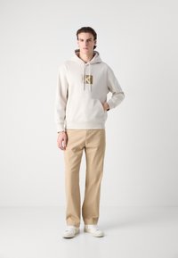 Hanorac bej deschis cu logo pe față, glugă cu șnur și buzunar tip cangur; asortat cu pantaloni bej deschis și adidași albi.