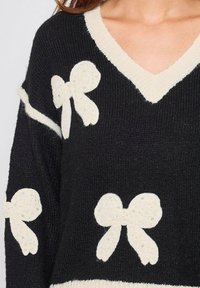 Pull noir avec des nœuds texturés de couleur crème ornés de perles, un décolleté en V et des détails côtelés au niveau de l'ourlet et des poignets.