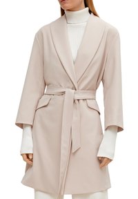 Hellrosa Wickeljacke mit einem Schalkragen, weiten Ärmeln und Bindegürtel. Verfügt über zwei Seitentaschen und wird über einem weißen Rollkragenpullover getragen.