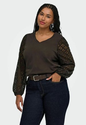 Kvinde med langt flettet hår iført en mørk V-hals top med gennemsigtige mønstrede lange ærmer og mørkeblå jeans, stående med den ene hånd i lommen.