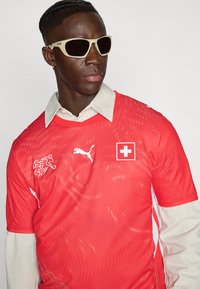 Vörös mez hullámos textúrával, fehér Puma logóval és svájci zászló folttal. Világos inggallérral és bézs napszemüveggel kísérve.