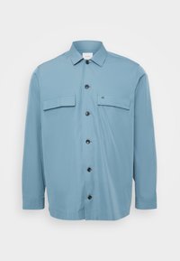 Camicia azzurra chiara con due taschini sul petto, bottoni neri e una texture liscia. Presenta un colletto e maniche lunghe.