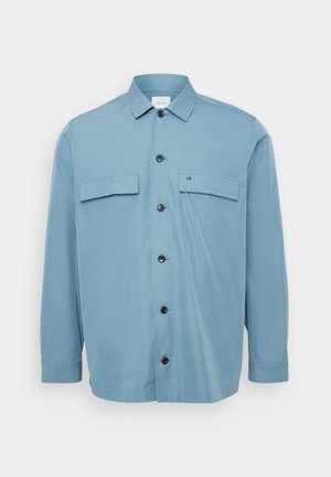 Svijetloplava košulja na gumb s dva džepa na prsima, crnim gumbima i glatk teksturom. Ima ovratnik i dugačke rukave.