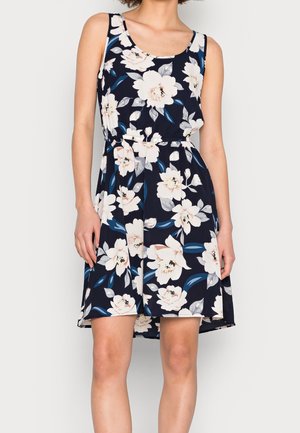 Ärmelloses marineblaues Kleid mit großem weißem und hellrosa Blumenmuster, tailliert und mit einem Saum oberhalb der Knie, an einer stehenden Person.