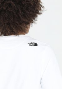 Person med lockigt hår som bär en vit skjorta med The North Face-logotypen på övre ryggen, vänd bort från kameran.