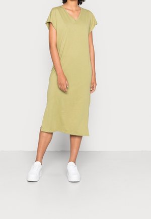 Femme debout portant une robe ample à manches courtes, longueur genou, de couleur vert olive, et des baskets blanches à plateforme sur un fond clair uni.