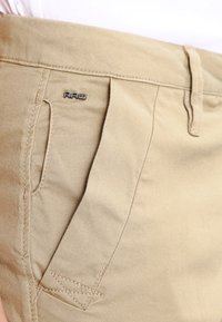 Close-up van een beige broekzak met een zichtbare riemlus en een klein metalen "RAW" logo boven de naad van de zak.