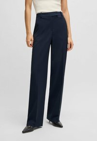 Pantaloni blu navy su misura con gamba dritta, realizzati in un tessuto testurizzato. Presentano passanti per cintura e una finitura liscia, abbinati a scarpe nere a punta.