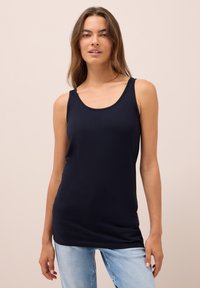 Una donna con lunghi capelli castani che indossa un top senza maniche nero e jeans azzurri chiari, in piedi su uno sfondo beige semplice.