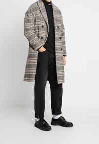 Cappotto a quadri grigio e nero con accenti rossi, design doppiopetto, indossato sopra una maglia nera e abbinato a jeans neri e scarpe.