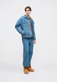 Jaqueta de ganga em azul claro, com colarinho, dois bolsos no peito, combinada com jeans azuis de corte solto e botas de camurça claras.