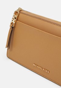 Pochette en cuir couleur tabac Michael Kors avec fermeture éclair dorée, breloque logo MK et détail de poche avant sur fond blanc uni.