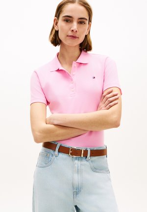Kvinde med kort brunt hår iført en pink polo-shirt og lyseblå jeans med et brunt bælte, armene krydset, stående mod en ensfarvet baggrund.