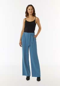Szerokie spodnie w niebieskim materiale o gładkiej fakturze, z paskiem w talii i bocznymi kieszeniami, zestawione z czarną koszulą na ramiączkach.