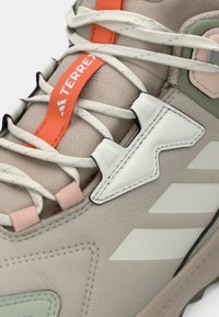 Αθλητικό παπούτσι σε μπεζ χρώμα με υφή υφάσματος, λευκές λεπτομέρειες, πορτοκαλί κορδόνια, πράσινα στοιχεία και εμφανές λογότυπο στην γλώσσα και στο σχέδιο της πλευράς.