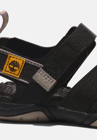Sandal preto em tecido com detalhes cinzentos, sola texturizada, tiras ajustáveis e um emblema amarelo. Design robusto com material respirável.