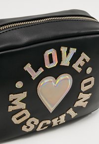 Musta nahkainen laukku kultaisella vetoketjulla, koristeltu holografisella sydämellä ja sanoin "LOVE MOSCHINO" kirjottuna.