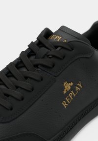 Black leather sneakers featuring a textured surface, round laces, a rubber sole, and gold logo detailing.