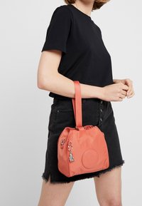 Kobieta w czarnym T-shircie i spódnicy, trzymająca na lewym ramieniu małą koralową torebkę z breloczkiem w kształcie klucza, na tle jednolitego tła.