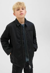 En blond kille iklädd en ofodrad svart jacka och mörka byxor med händerna i fickorna, vänd framåt mot en ljus enkel bakgrund.