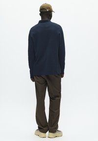 Jūras spēku tumši zils garām piedurknēm polo krekls ar apkakli, apvienots ar brūniem biksēm un bēšām kurpēm. Modelis valkā khaki cepuri un ir pagriezies prom no kameras.