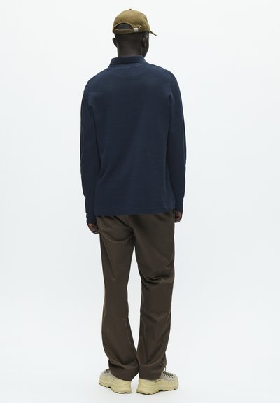 Polo a maniche lunghe blu navy con colletto, abbinato a pantaloni marroni e scarpe beige. Il modello indossa un cappello kaki e guarda lontano dalla fotocamera.