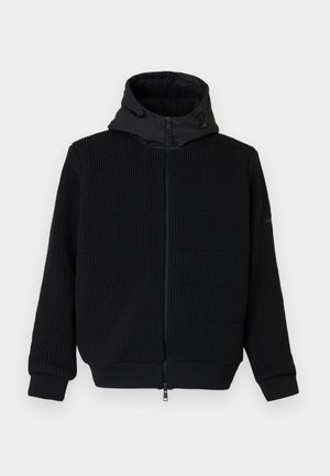 Musthave musta kapuutsiga, millel on soonikpind, sile kapuuts ja nöör. Omab pikki varrukaid ja elastseid puusaosa. Ees on tõmblukuga sulgemine.