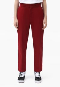 Červené zúžené kalhoty vyrobené z hladkého bavlněného materiálu s poutky na opasek, doplněné černými teniskami s bílými šňůrkami a podrážkami.