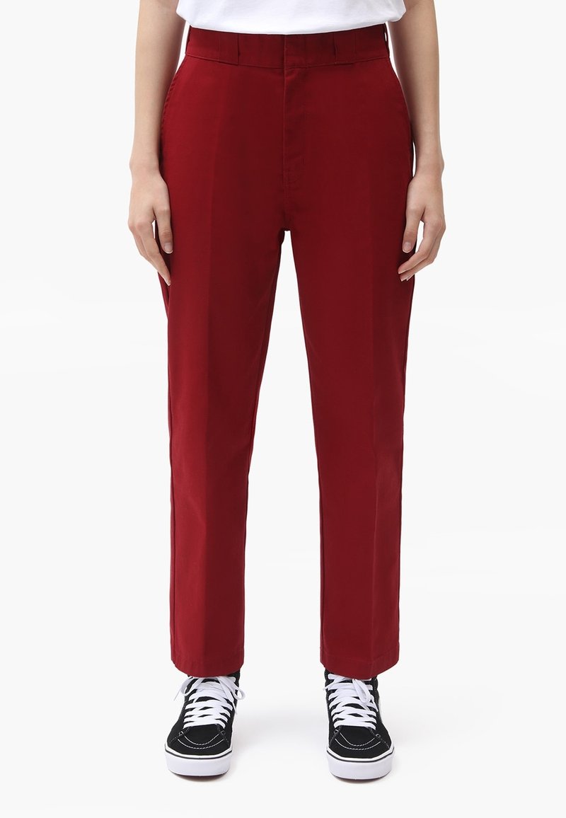 Červené zúžené kalhoty vyrobené z hladkého bavlněného materiálu s poutky na opasek, doplněné černými teniskami s bílými šňůrkami a podrážkami.