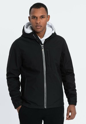 Veste à capuche noire avec une fermeture éclair à l'avant, des poignets élastiques et une poche sur la poitrine, superposée à un sweat-shirt gris. Texture lisse et design minimaliste.