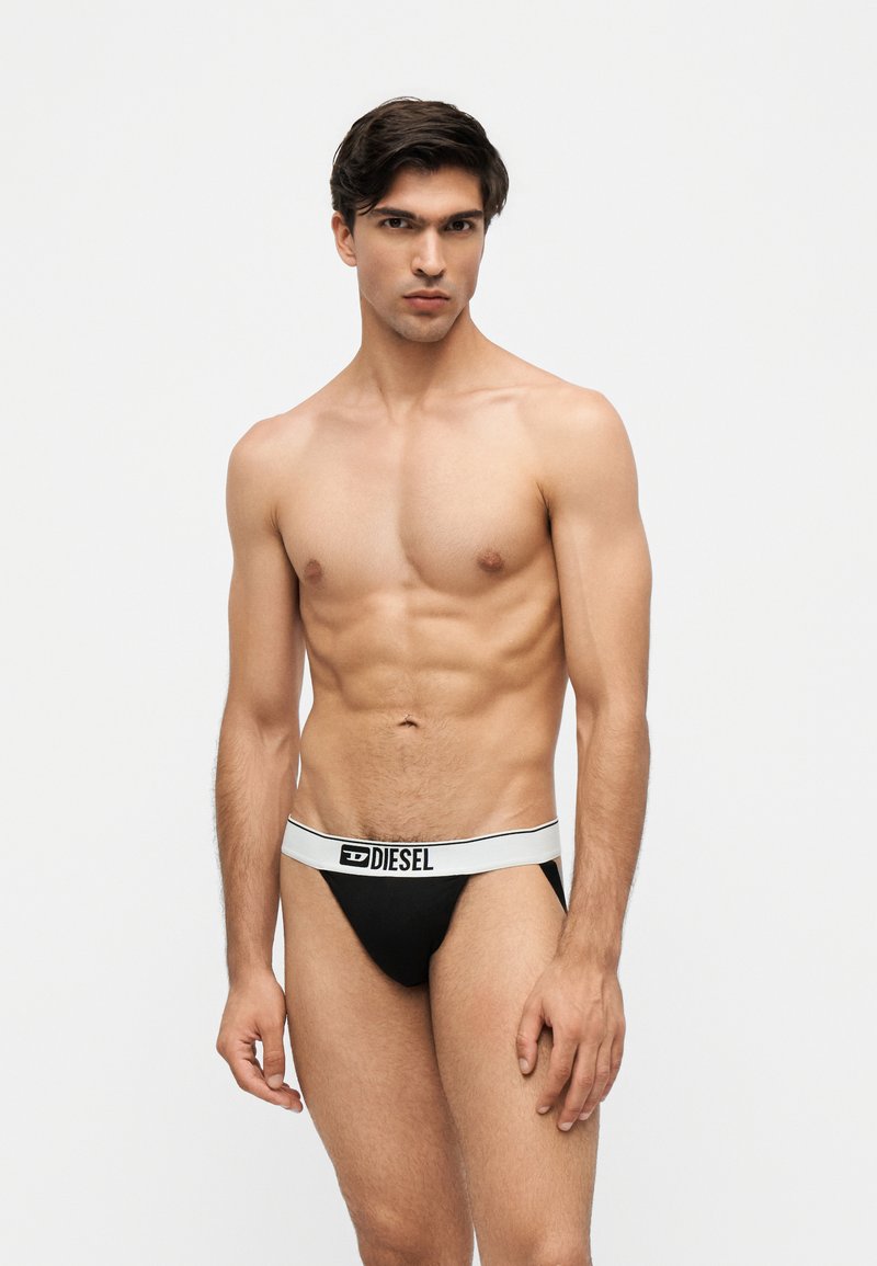Homme musclé aux cheveux foncés portant un sous-vêtement Diesel noir avec une ceinture blanche, debout devant un fond clair uni.