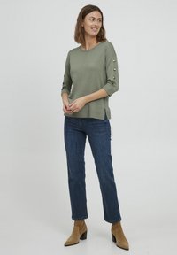 Haut en maille vert olive avec des manches trois-quarts et des fentes latérales, présentant des accents de bouton sur les manches, associé à un jean droit bleu foncé et des bottines beige.