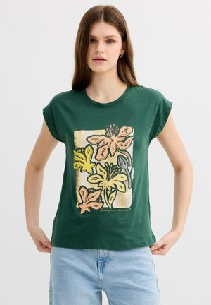 Joven mujer con camiseta verde con gráfico floral pastel sostiene el dobladillo, combinada con jeans denim azul claro sobre fondo liso.
