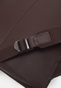 Sac en cuir marron foncé avec bandoulière en tissu assorti et boucle métallique argentée, présenté en gros plan sur fond blanc.