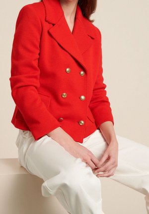 Punainen kaksirivinen bleiseri kultaisilla napeilla, jossa on lovikaulus ja teksturoitu kangas, yhdistettynä valkoisiin housuihin, istuu neutraalilla pinnalla.