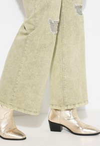 Bottes de cheville métalliques dorées avec un bout pointu et un dessus texturé, associées à un jean large vert clair avec des détails effilochés au niveau des genoux.