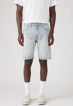 Homme portant un short en jean bleu clair avec un t-shirt blanc, des chaussettes blanches et des baskets en toile beige, debout devant un fond uni.