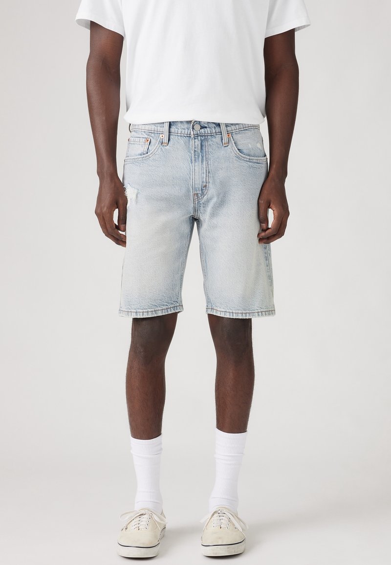 Homme portant un short en jean bleu clair avec un t-shirt blanc, des chaussettes blanches et des baskets en toile beige, debout devant un fond uni.