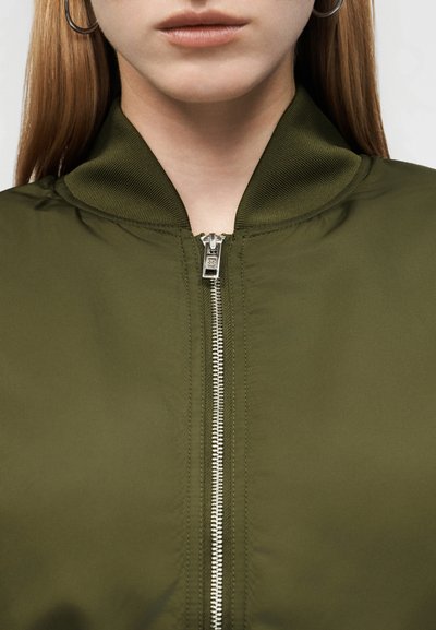 Γυναίκα που φοράει χακί μπουφάν bomber φερμουάρ κλειστό μέχρι τον γιακά, με ασημένια κρίκους σκουλαρίκια και ίσια, ανοιχτόχρωμα καστανά μαλλιά.