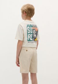 T-shirt beige a maniche corte con una stampa grafica colorata sulla parte posteriore, abbinata a pantaloni corti beige chiaro con una tasca posteriore con un accento dorato.