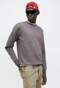 Homme portant une casquette rouge à l'envers, des lunettes de soleil noires, un sweat-shirt gris et un pantalon beige, debout avec une main dans la poche devant un fond uni.