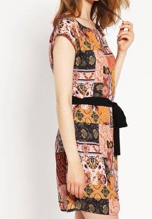 Kobieta ubrana w sukienkę w krótkim rękawem o wzorze patchworkowym w odcieniach pomarańczu, różu i czerni, z czarnym paskiem w talii, stojąca bokiem.