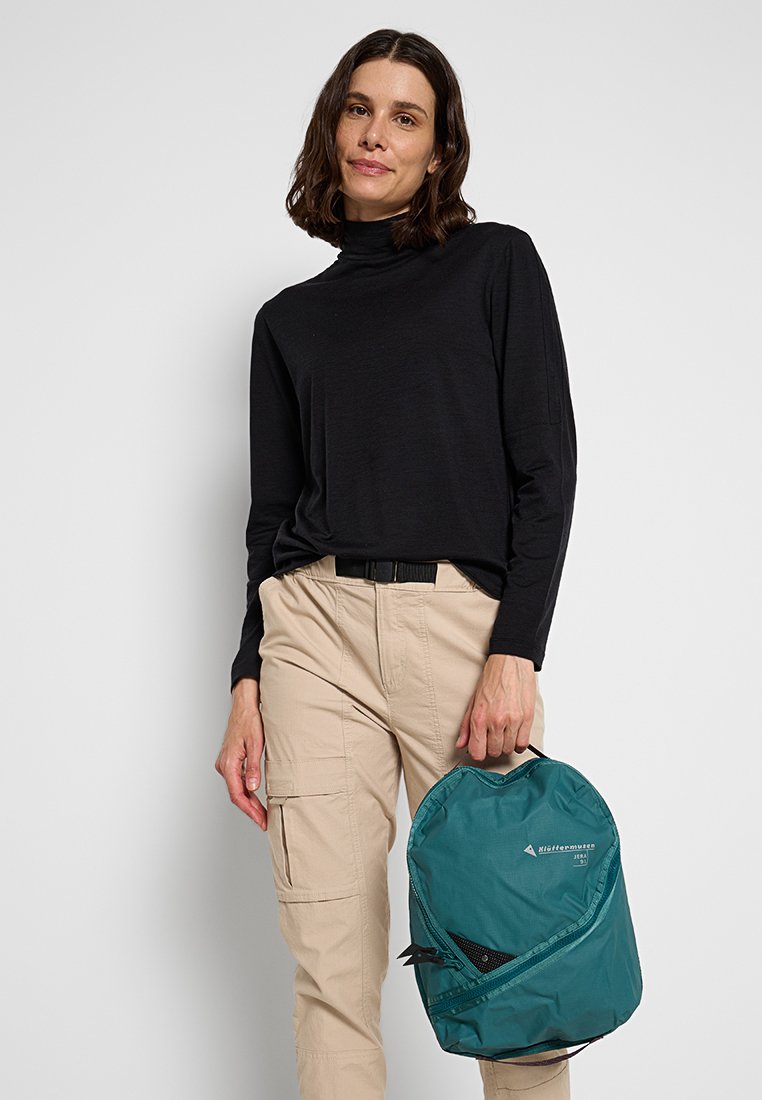 Frau trägt ein schwarzes Langarmoberteil und beigefarbene Cargo-Hosen, hält einen türkisen Rucksack am Griff vor einem schlichten Hintergrund.