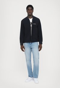 Marineblå bomberjakke med lynlås, ribbede manchetter og to lommer over en hvid T-shirt med grafik, parret med lyseblå denim og hvide sneakers.